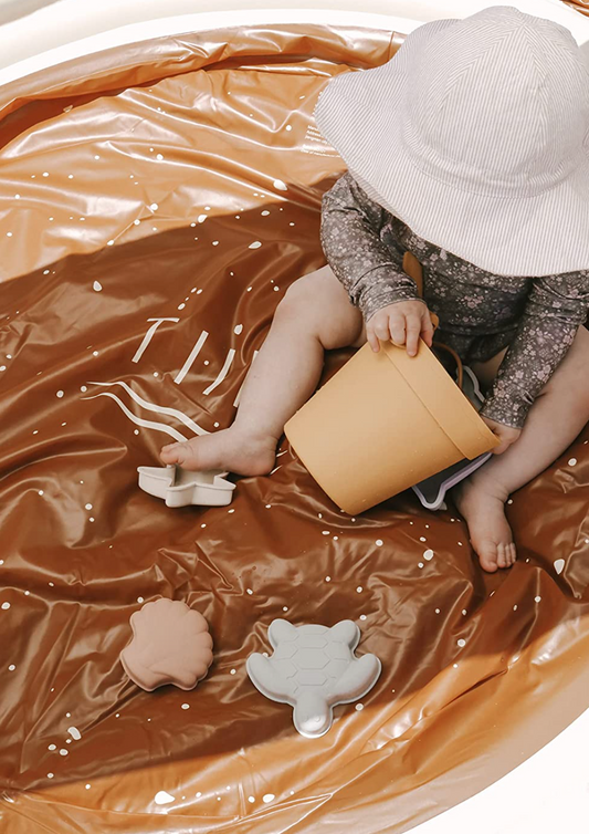 Overhead view of a toddler in a wide-brim sun hat sitting in the Tiipikids Beach Pool, holding a tan bucket, with pale silicone sand moulds floating in the shallow water
