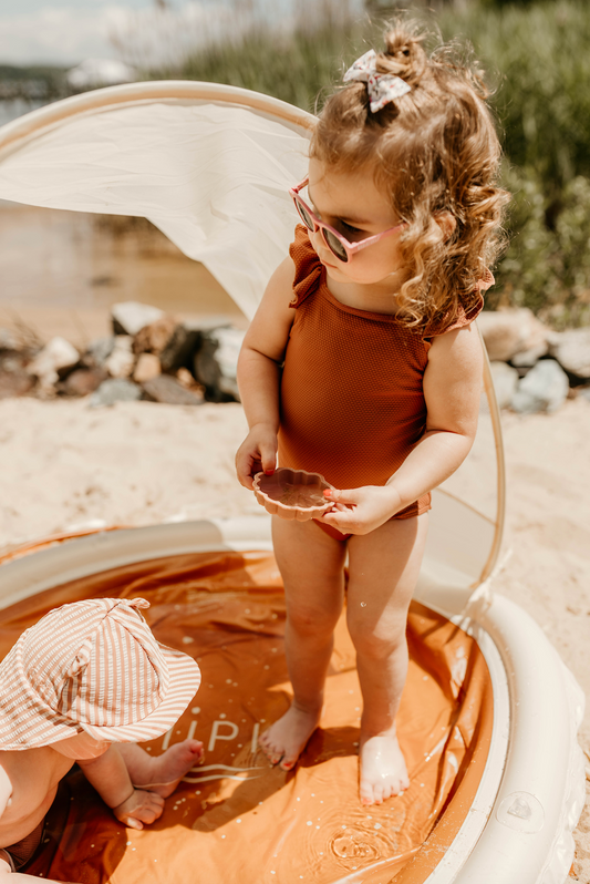 A girl in a rust swimsuit and sunglasses standing at the edge of the Tiipikids Beach Pool while a younger child in a sun hat sits in the water beside her
