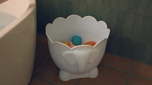 Bath Buddy, an off-white organizer with a scalloped rim holding small colorful toys, standing on a floor beside a bathtub