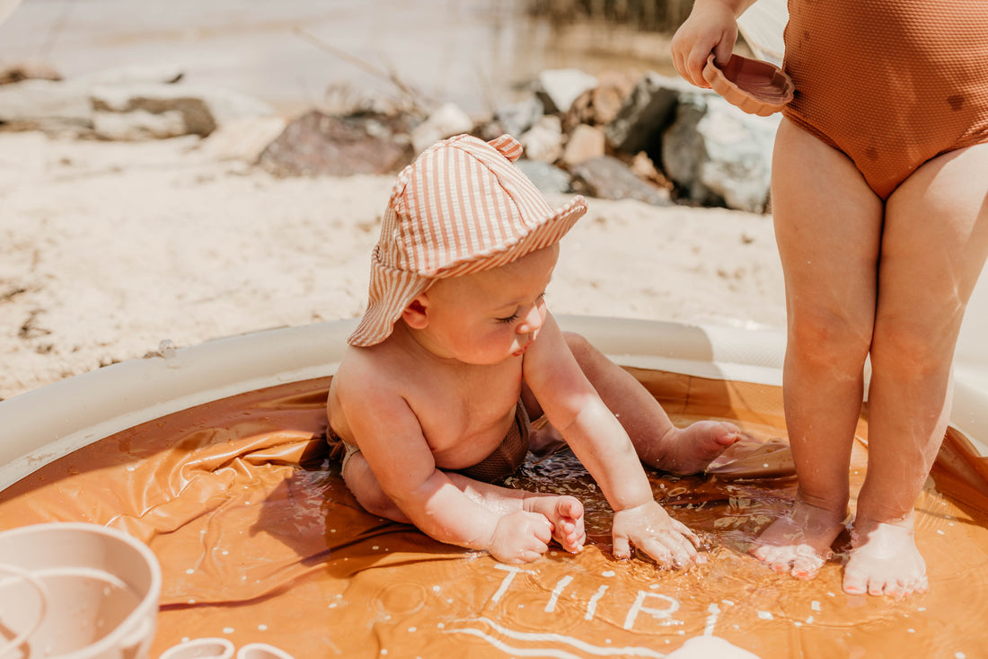 A baby in a sun hat sitting in the water-filled Tiipikids Beach Pool on the shoreline, an adult crouching alongside and rocks in the background