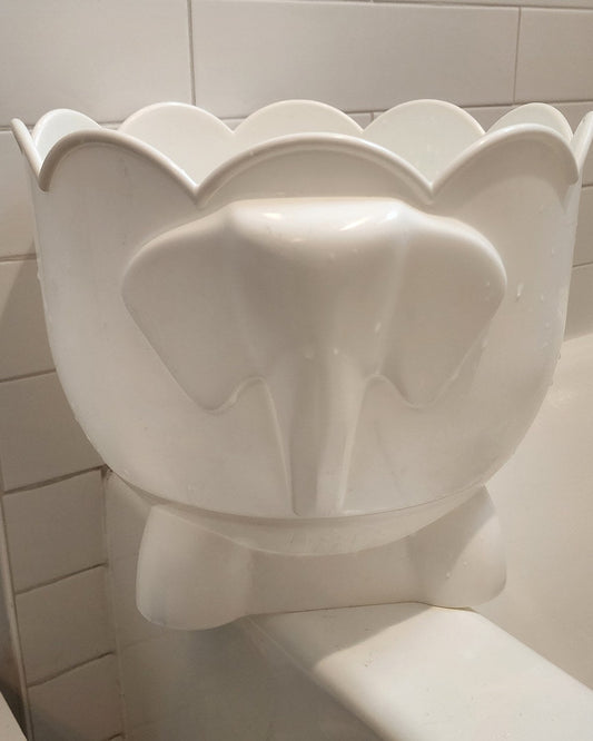 Bath Buddy, a white plastic drum with a scalloped rim and a raised relief on its front, standing against a white tiled wall