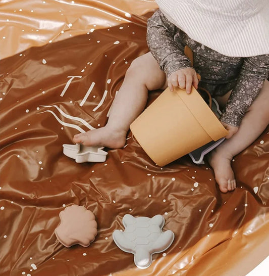 A child sitting outdoors holding a beige beach bucket with a shovel, beach toys scattered around.